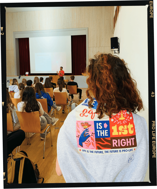 A photo of Benjamin Famula giving a LifeTALK, with Karolina at the back of the room wearing a grey ProLife Europe sweater featuring the phrase "the right to life is the 1st right"