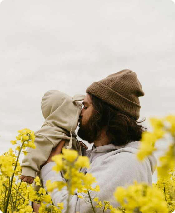 A father wearing a beanie holds up his young child closely to his face, and they're surrounded by yellow flowers