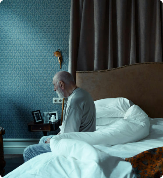 Photo of an elderly man sitting pensively and hunched over at the edge of the bed