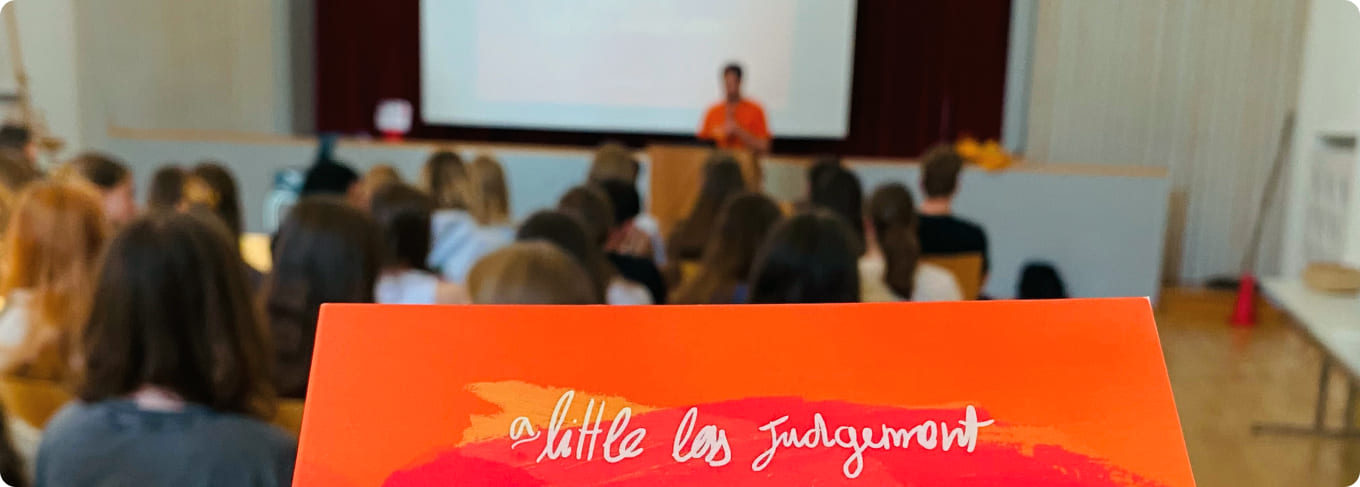 A photo of a ProLife Europe member giving a LifeTALK in an open hall to a seated audience. In the foreground there is an orange flyer with the phrase "a little less judgement, a little more truth"