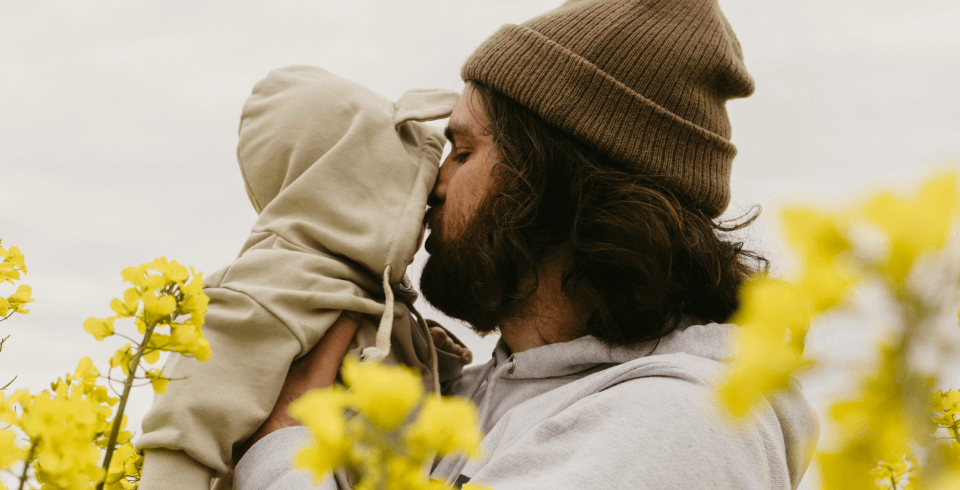 A father wearing a beanie holds up his young child, and they're surrounded by yellow flowers