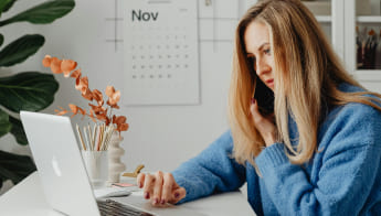 A photo of a young blonde woman working on a laptop and talking on her mobile phone at the same time