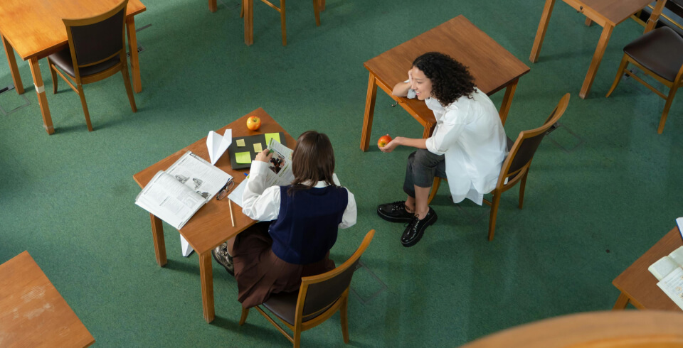 Two female school students sit at their desks and talk to each other, smiling