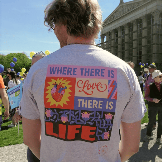 A man showing the back of a grey ProLife Europe t-shirt, featuring the phrase "where there is love there is life"