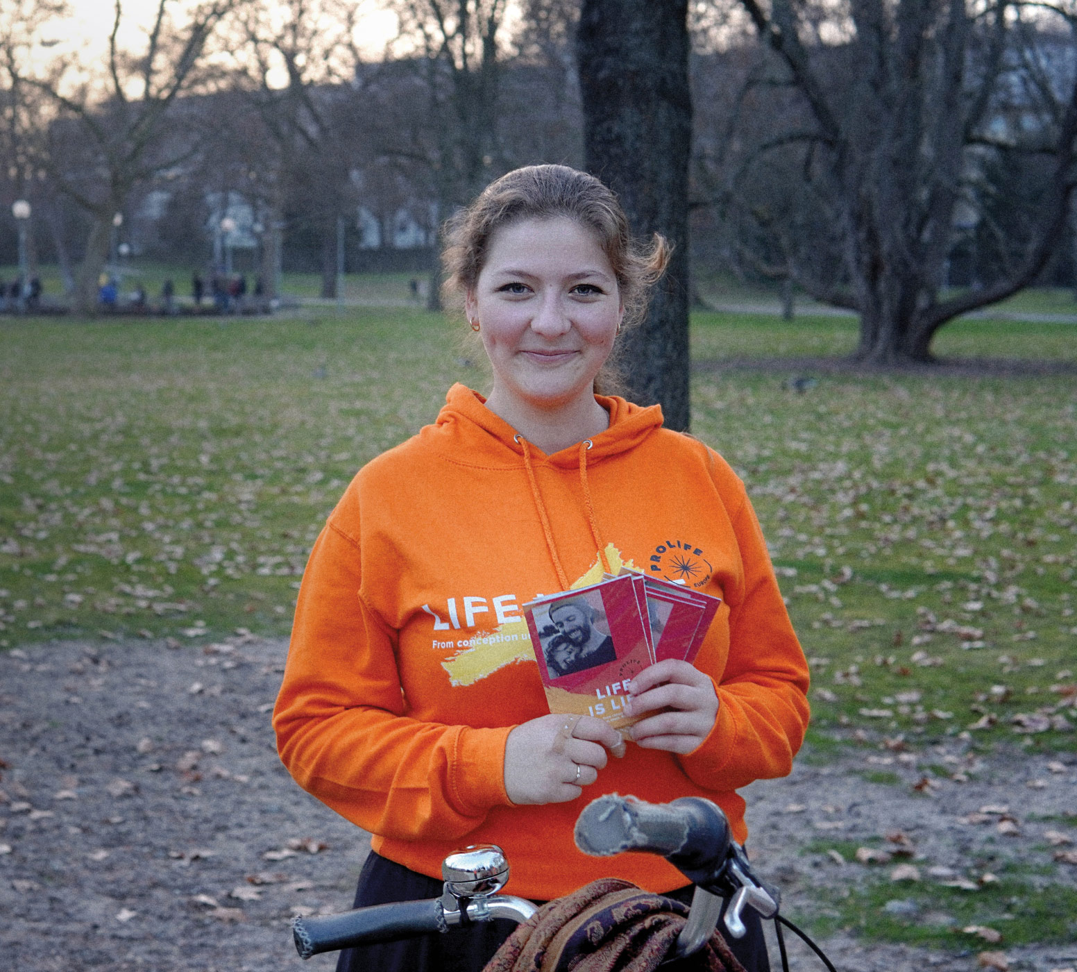 A lady from the ProLife Europe team holding flyers and wearing an orange 'Life is life' hoodie, in an outdoors park environment