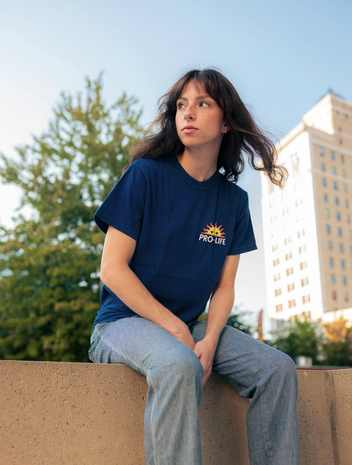 A young woman wearing the navy t-shirt from the ProLife Europe merchandise collection,  sitting on a concrete wall in an urban setting